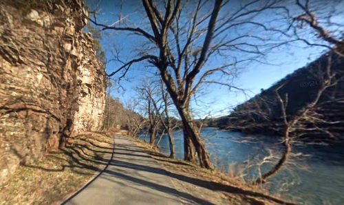 A winding path alongside a river, framed by bare trees and rocky cliffs under a clear blue sky.