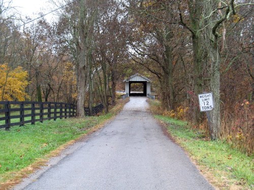 A gravel road leads to a covered bridge, surrounded by trees with autumn foliage and a weight limit sign.
