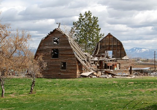 A dilapidated barn with a collapsed roof, surrounded by grass and trees, against a backdrop of cloudy mountains.