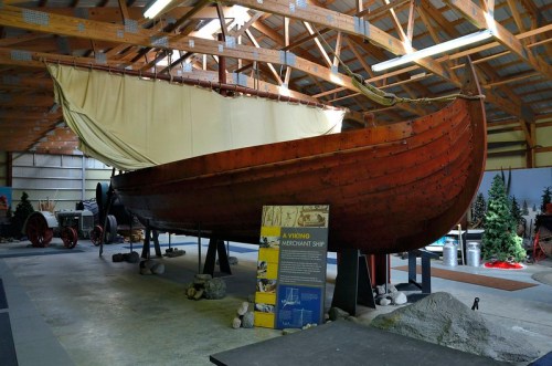 A wooden Viking merchant ship displayed indoors, with a sail and informational signage nearby.