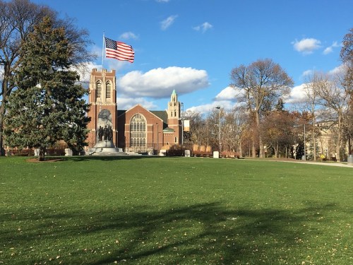A grassy area with a flagpole, featuring a church and clock tower against a blue sky with scattered clouds.