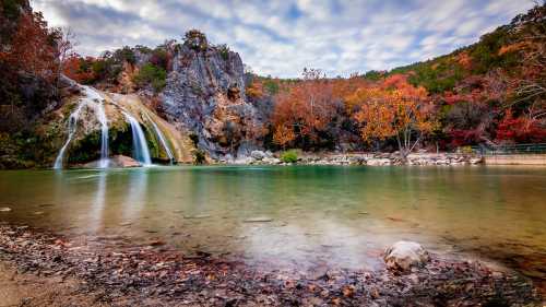 A serene waterfall cascades into a calm pool, surrounded by autumn foliage and rocky cliffs under a cloudy sky.