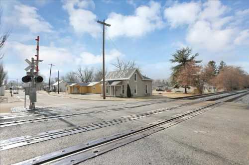 A quiet street scene featuring a railroad crossing, a house, and a few trees under a partly cloudy sky.