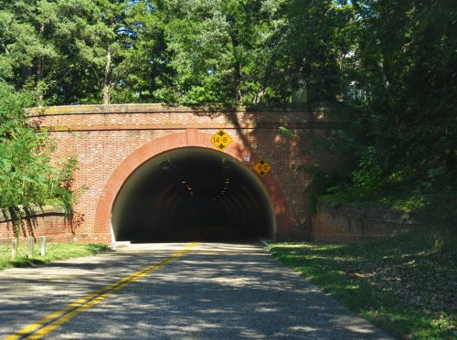 A brick tunnel entrance with height and width warning signs, surrounded by trees and a clear blue sky.