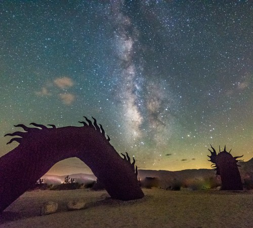 A starry night sky with the Milky Way above two abstract sculptures resembling arches in a desert landscape.