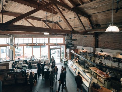 A bustling market interior with wooden beams, people mingling, and various food displays in a bright, open space.