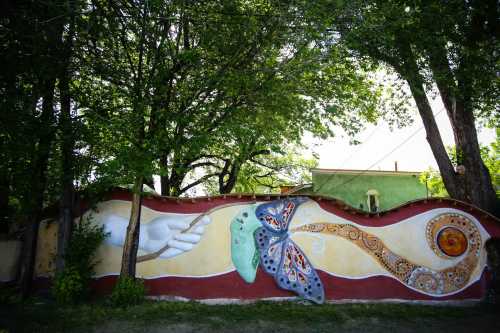 A colorful mural featuring a butterfly, abstract shapes, and a hand, surrounded by lush green trees.