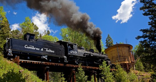 A vintage steam train emits black smoke while passing a wooden water tower in a lush, green forest.
