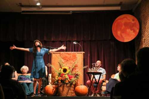 A speaker with arms outstretched stands at a podium, surrounded by an audience and autumn decorations, including pumpkins.