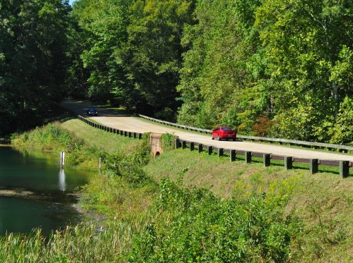 A scenic road curves alongside a calm body of water, surrounded by lush green trees. Two cars are visible on the road.