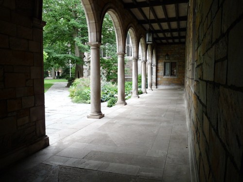 A stone corridor with arches, leading to a green garden area, surrounded by trees and a peaceful atmosphere.