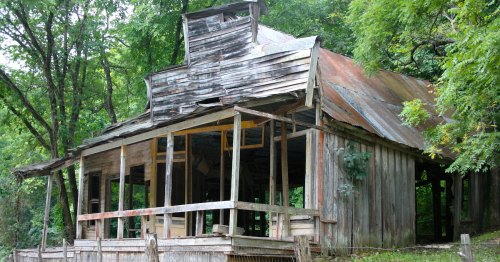 An old, weathered wooden building with a rusty roof, surrounded by trees and overgrown vegetation.