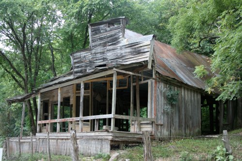 A weathered, abandoned wooden building with a rusty roof, surrounded by lush greenery and a wooden fence.