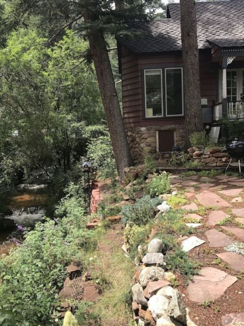A cozy house surrounded by trees and a stone pathway, with a stream flowing nearby.