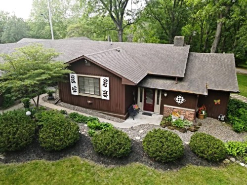 A cozy brown house surrounded by greenery and decorative landscaping, featuring unique shutters and a welcoming entrance.