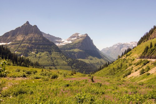 A scenic view of mountains and valleys, featuring lush greenery and patches of snow under a clear blue sky.