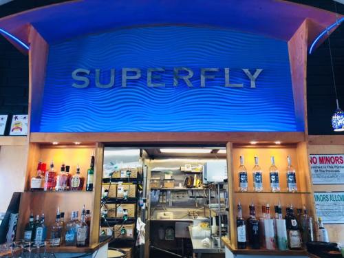 A bar area with the sign "SUPERFLY" illuminated in blue, showcasing various bottles of liquor and a kitchen in the background.