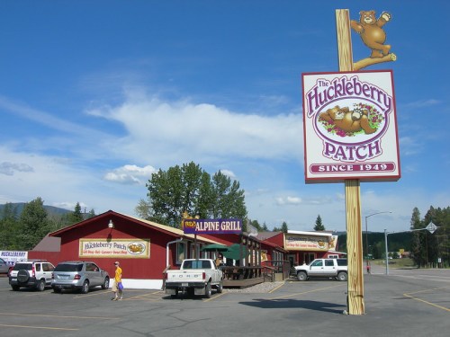 A colorful sign for "The Huckleberry Patch" restaurant, with a rustic building and parked cars in a scenic outdoor setting.