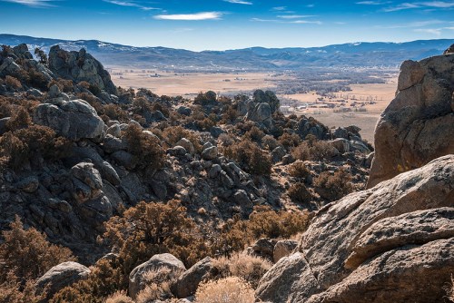 A panoramic view of rocky terrain overlooking a valley with distant mountains and a clear blue sky.