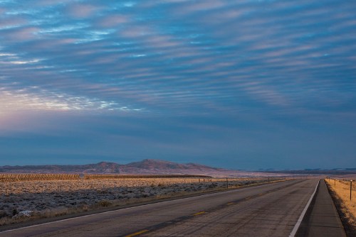 A wide, empty road stretches into the distance, flanked by open fields and mountains under a cloudy sky.