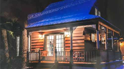 A cozy wooden cabin at night, illuminated by warm light, with a blue roof and a welcoming porch.