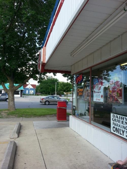 A storefront with an "Open" sign, featuring a menu display and a red newspaper stand outside. Trees in the background.