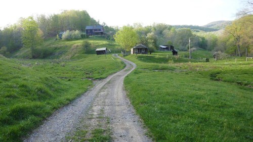 A gravel road winds through a green landscape with hills, trees, and rustic cabins in the distance.