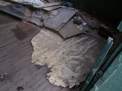 An abandoned room with peeling floorboards and scattered debris, including old cardboard and dirt.