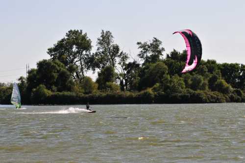 A kitesurfer rides the waves near a lush green shoreline, with another windsurfer in the background.