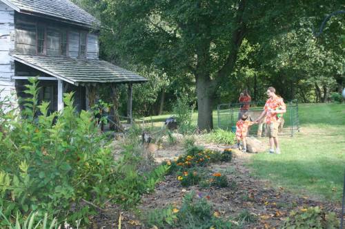 A group of people in colorful shirts working in a garden near an old house, surrounded by greenery and flowers.