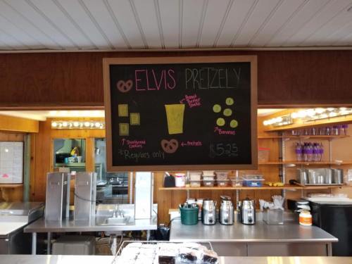 Chalkboard menu displaying "Elvis Pretzely" with images of pretzels, a drink, and prices in a cozy café setting.