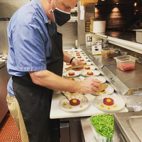 A chef in a mask plates dishes with colorful garnishes in a restaurant kitchen.