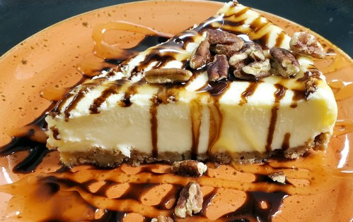 A slice of cheesecake topped with chocolate drizzle and pecans, served on an orange plate.