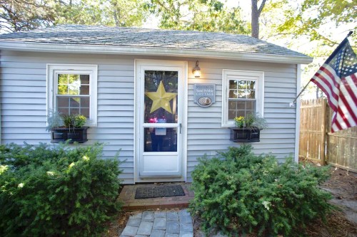 A small gray cottage with a star decoration on the door, surrounded by greenery and an American flag.