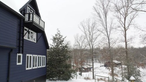 A snowy landscape featuring a blue house, trees, and distant barns in a serene winter setting.