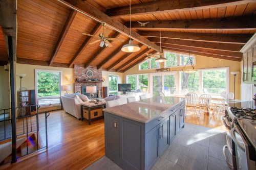 Bright, open living space with wooden beams, a stone fireplace, and a kitchen island, surrounded by large windows.