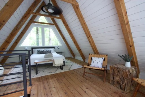 Cozy A-frame cabin interior with wooden beams, a bed, and a rustic chair, surrounded by nature.