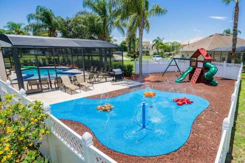 A sunny backyard with a pool, splash pad, playground, and seating area surrounded by palm trees and a white fence.