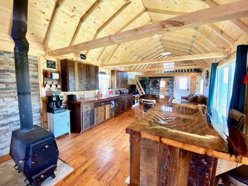 Cozy cabin interior with wooden beams, a stone fireplace, kitchen, and a rustic bar area. Bright and inviting atmosphere.