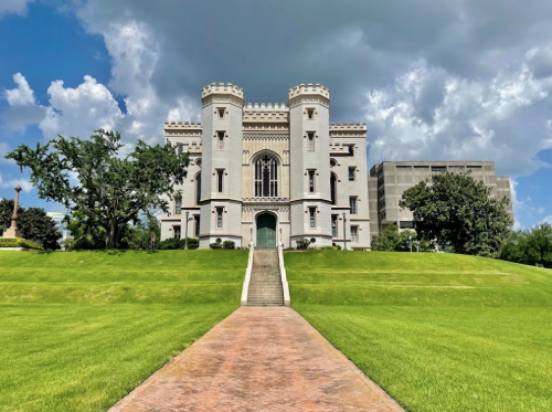 A historic castle-like building with towers, surrounded by green grass and trees, under a cloudy blue sky.