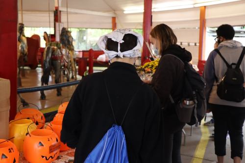 A group of people browsing a festive market with Halloween decorations and a carousel in the background.