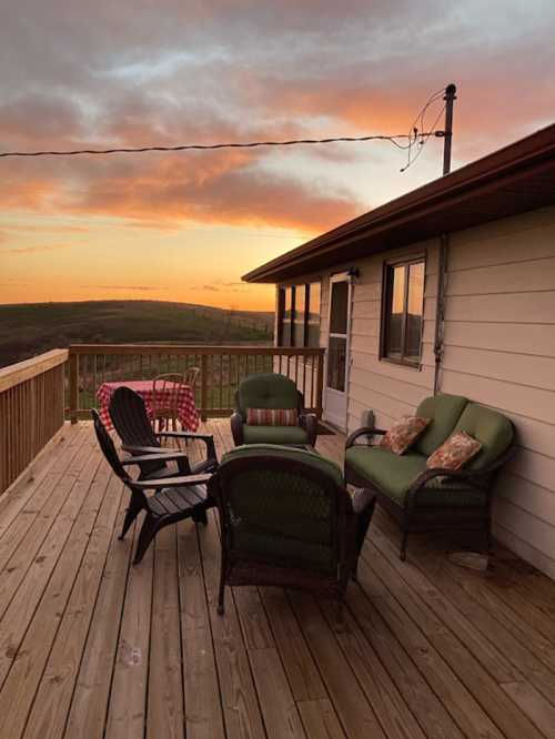 A cozy deck with green chairs and a table, overlooking a sunset sky with clouds and rolling hills in the background.