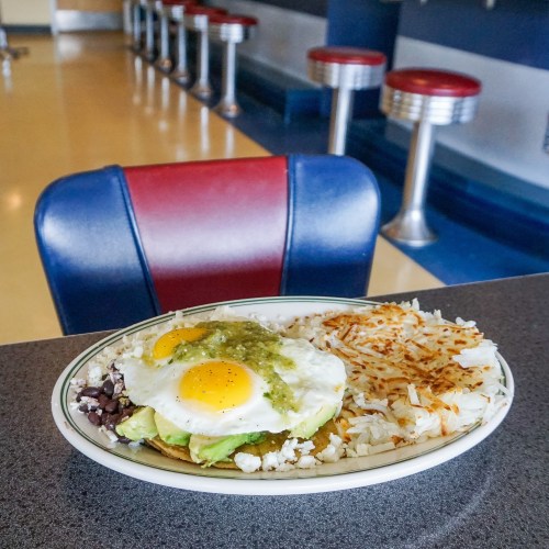 A plate of huevos rancheros with two eggs, avocado, salsa, and hash browns, set on a diner counter.