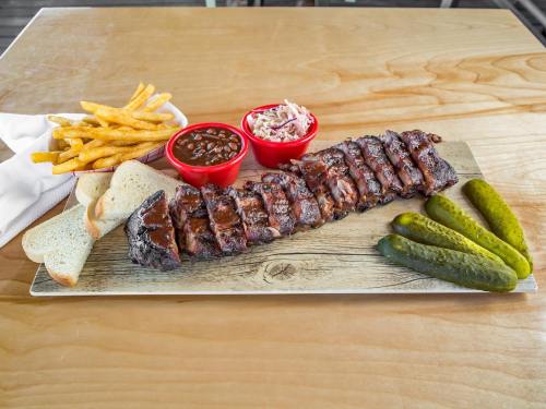 A wooden platter with BBQ ribs, fries, coleslaw, baked beans, pickles, and slices of bread.