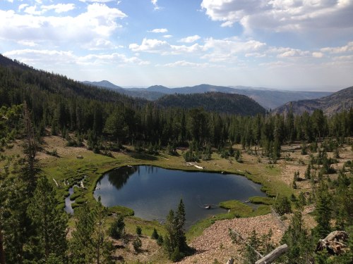A serene mountain landscape featuring a calm pond surrounded by lush greenery and distant mountains under a blue sky.