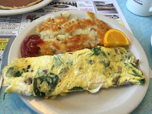 A plate with a spinach and mushroom omelet, hash browns with ketchup, and a slice of orange.