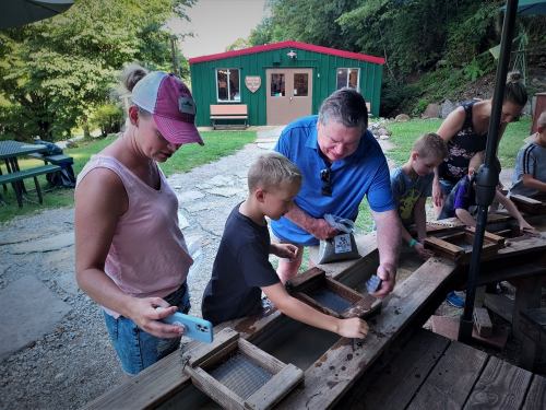 A family enjoys panning for gemstones at an outdoor activity area, with a green building in the background.