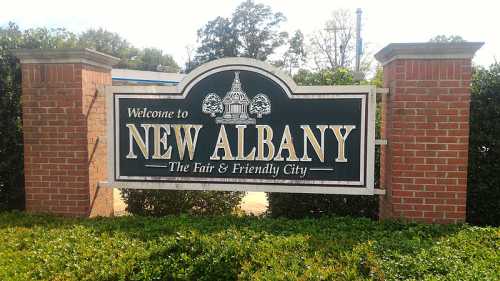 Sign welcoming visitors to New Albany, labeled "The Fair & Friendly City," surrounded by greenery.