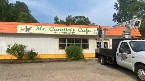 A worker on a lift adjusts the sign of Ms. Candia's Cafe, a white building with an orange roof and green lettering.