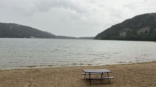 A picnic table on a sandy beach by a calm lake, surrounded by hills under a cloudy sky.